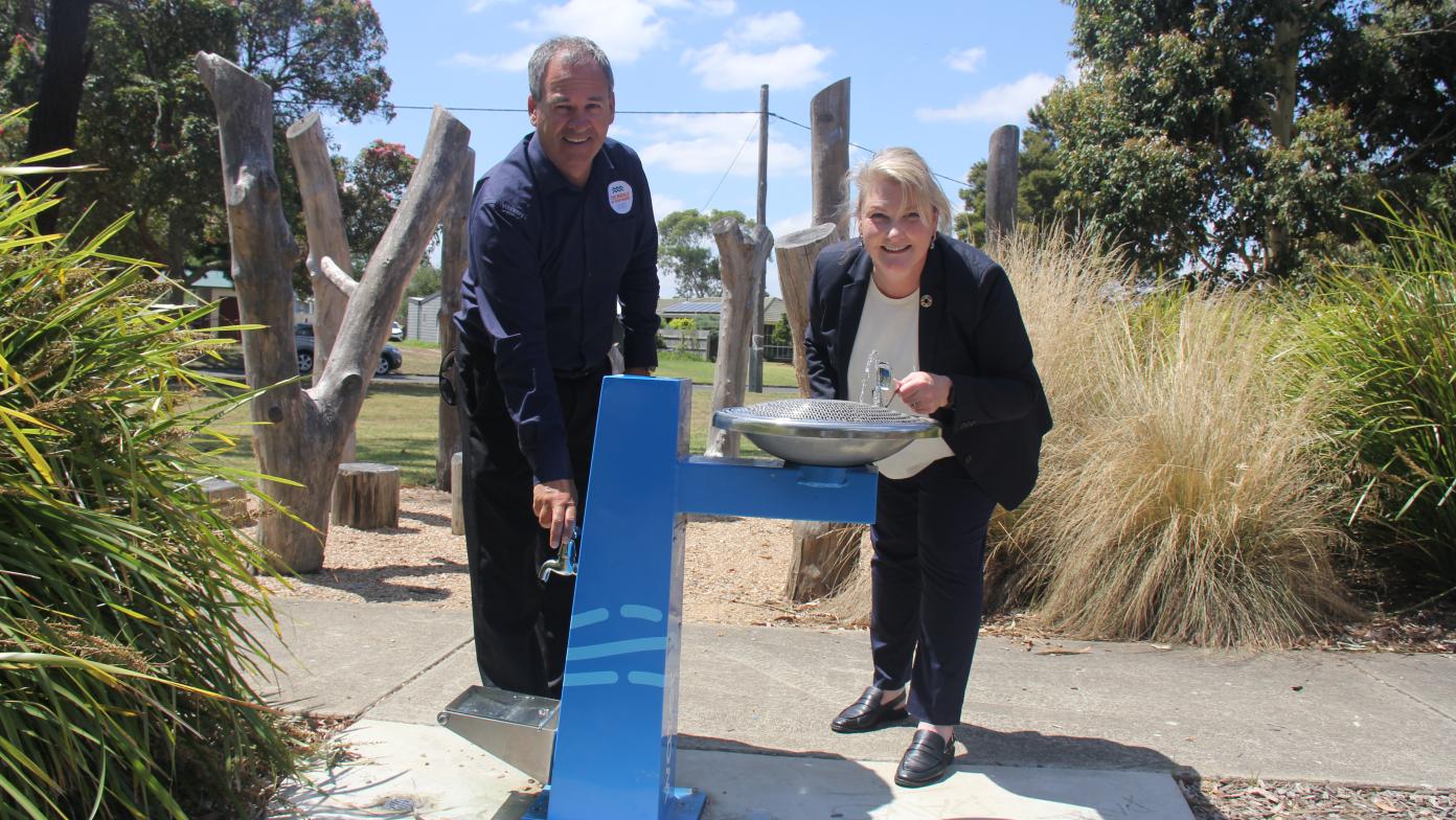 Man and woman stand at blue drinking fountain surrounded by shrubbery.