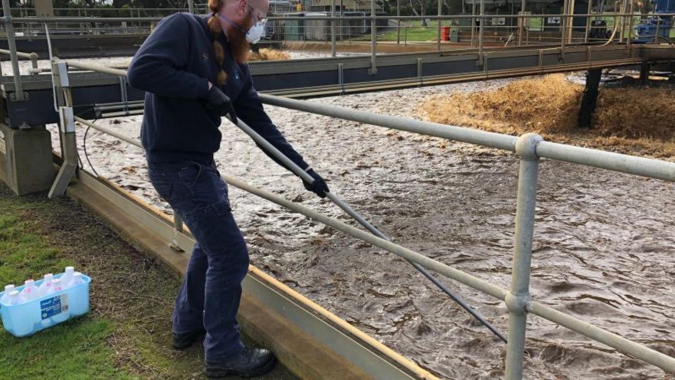 Man in Gippsland Water uniform uses a pole to test water. He wears a mask.