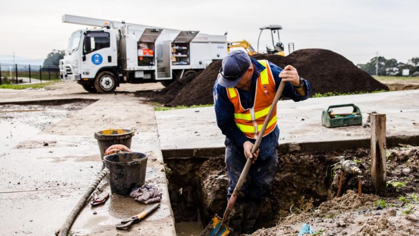 Man up to his knees in dirt digs a hole with shovel. He is in Gippsland Water uniform.