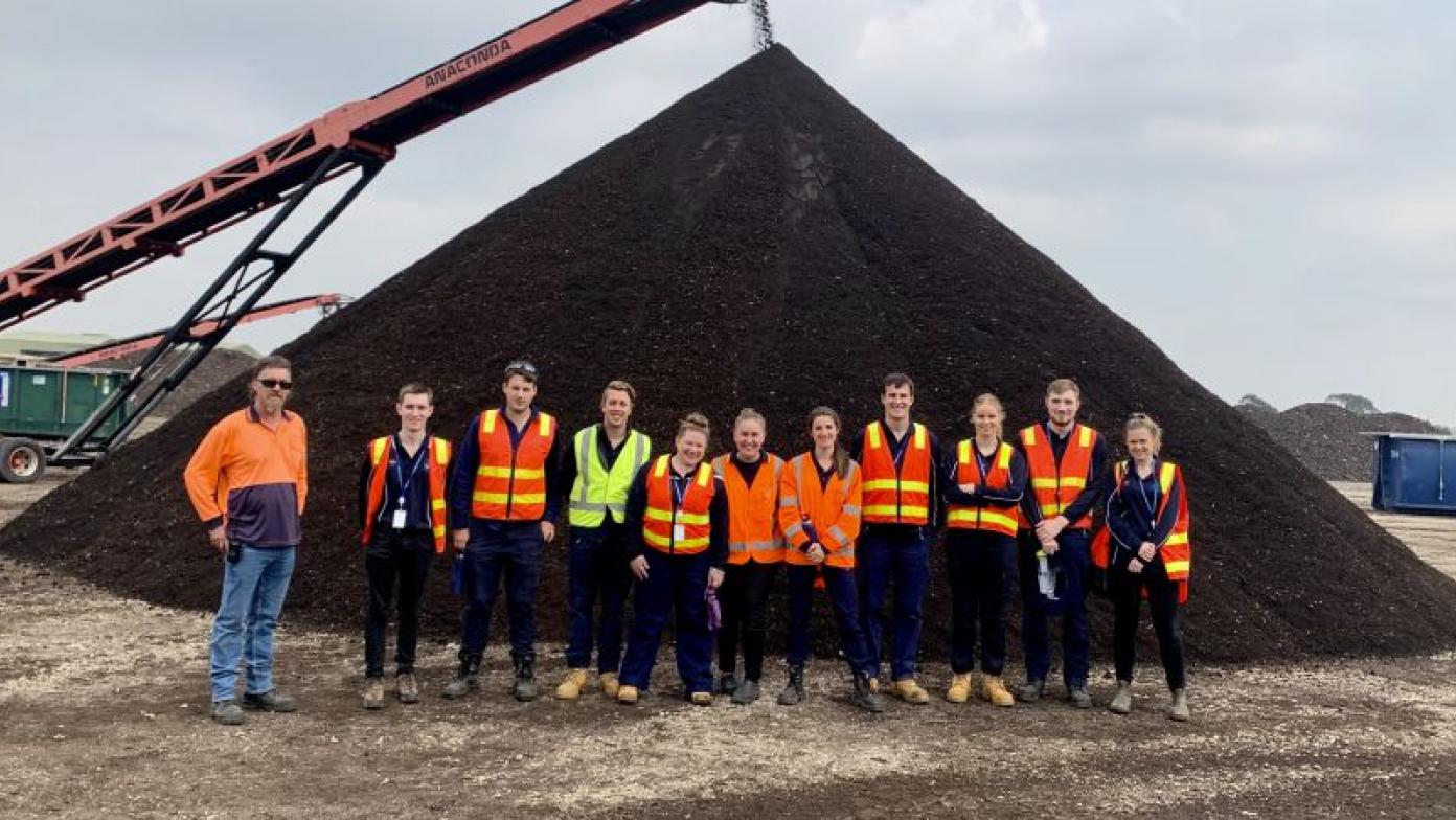 Eleven people in high visibility jackets stand in front of pie of dirt. They are smiling. 