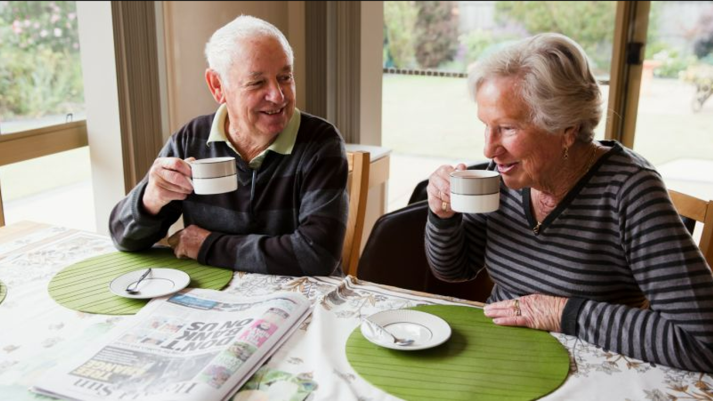 Elderly man and woman drink cups of tea at a dinner table. They are smiling.