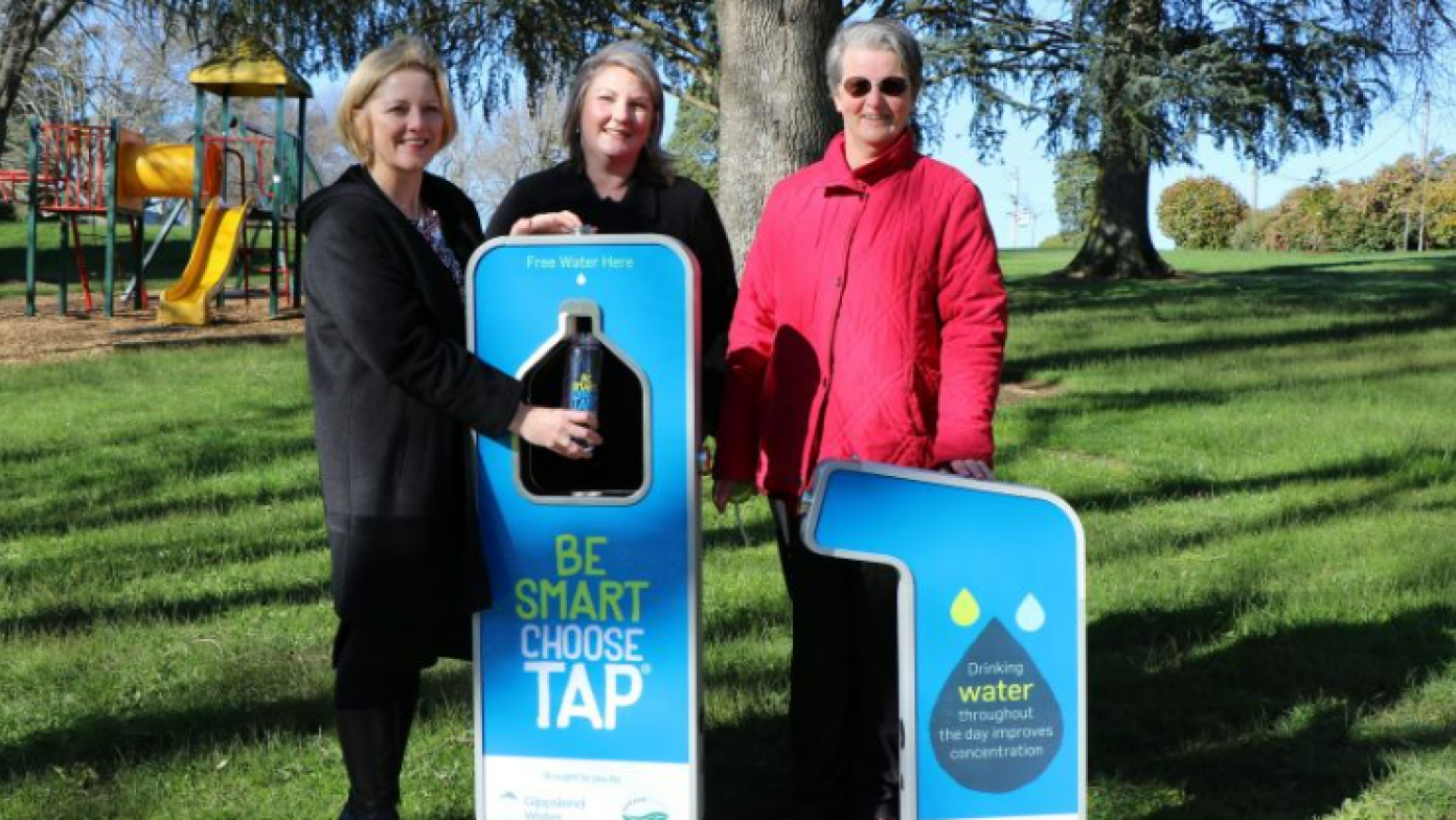 Three women stand behind water fountains in a park. 