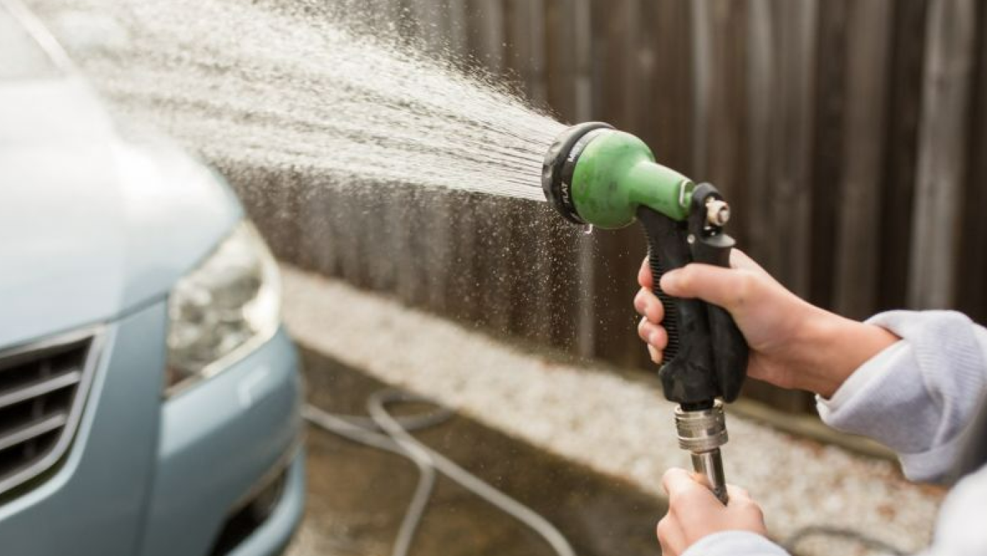 Water is being sprayed out of a hose at a light blue car. A person is holding the hose. 