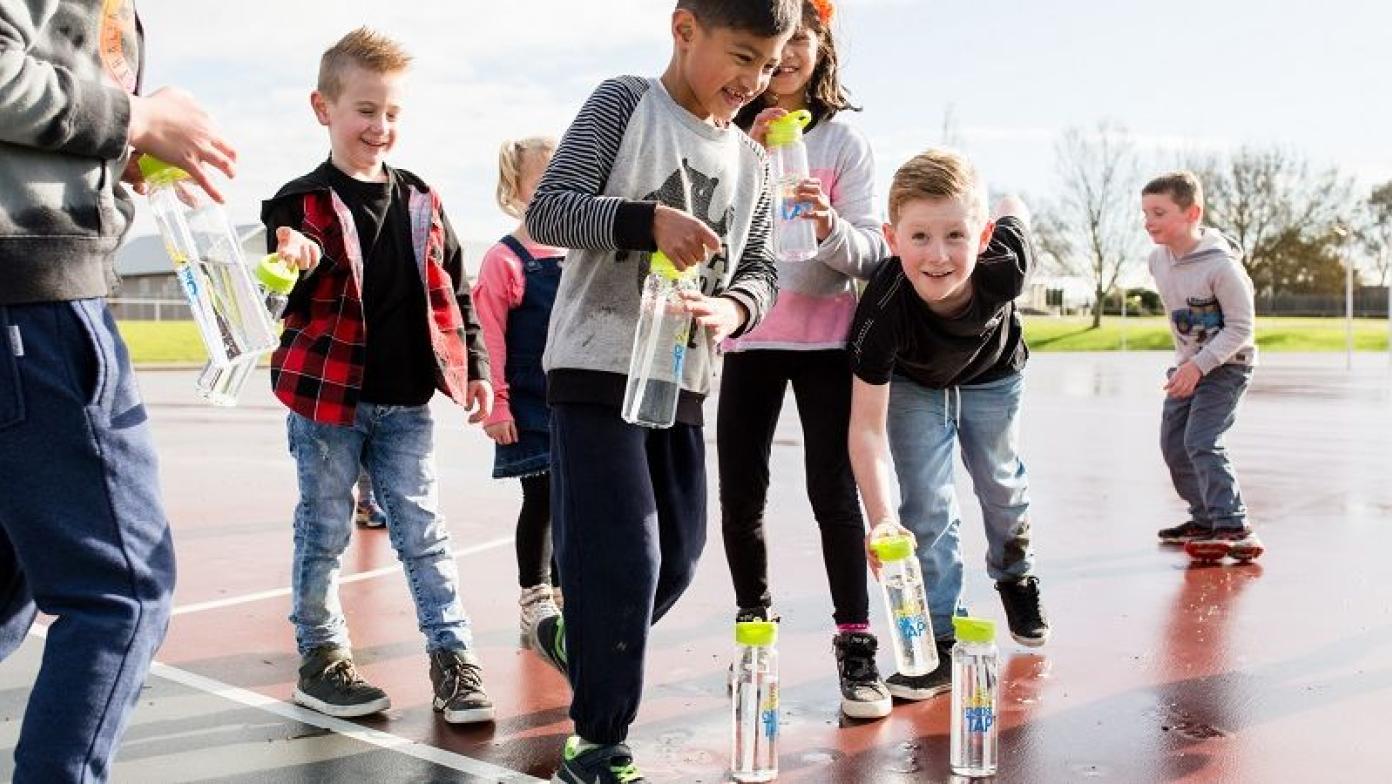 Children pick up water bottles off the ground of an orange netball court.