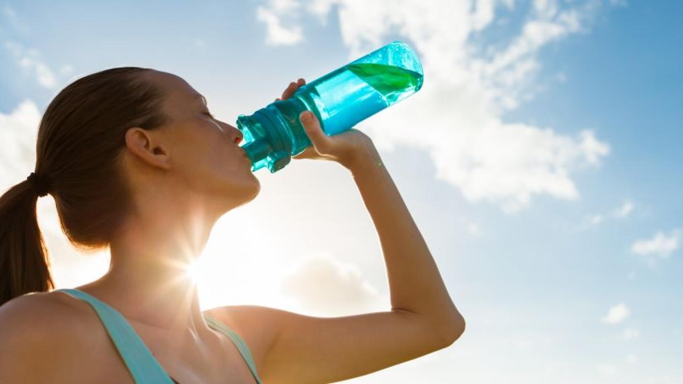 Sun shines behind woman drinking from a blue bottle of water.