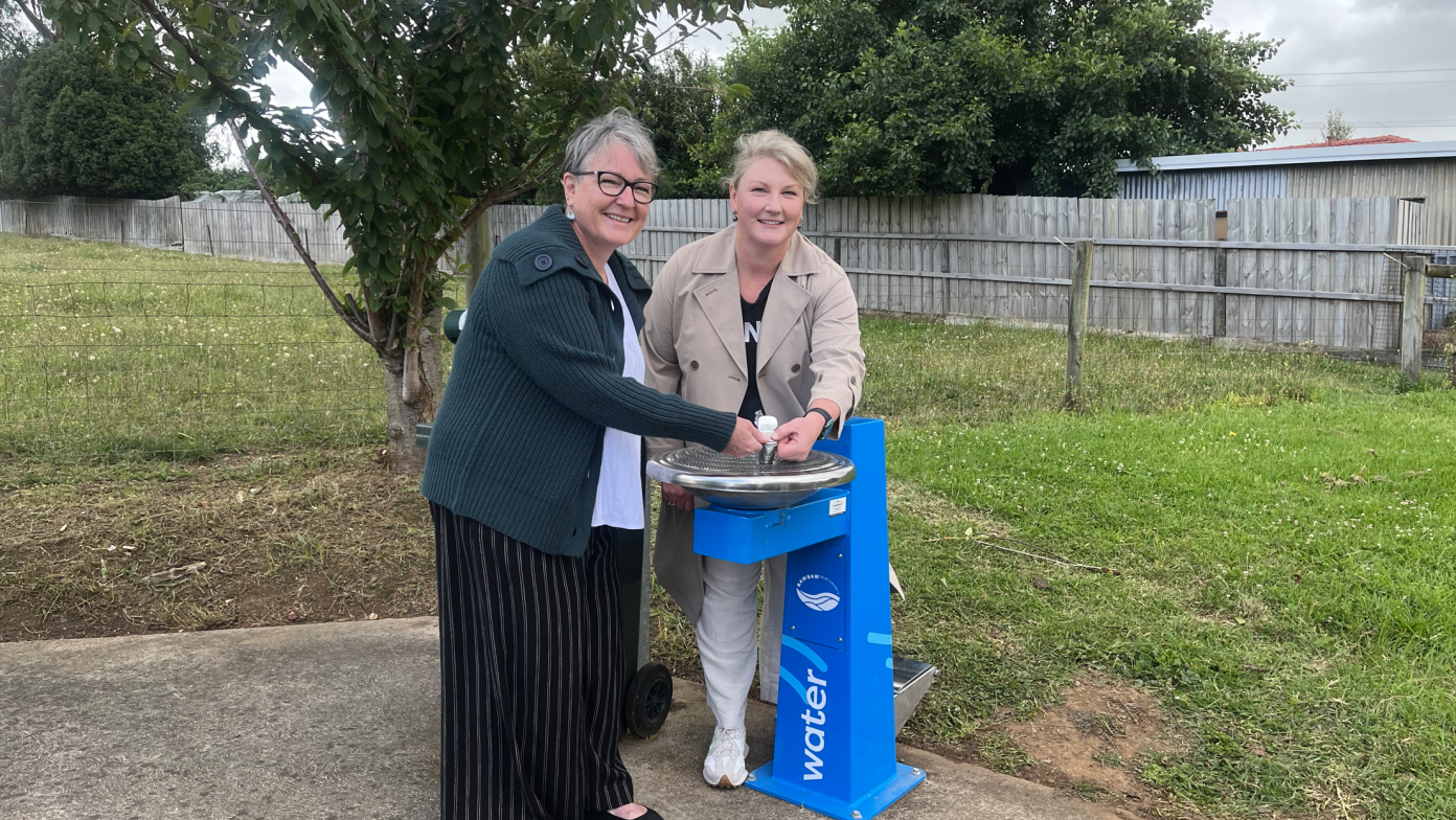 Two women stand behind a blue drinking fountain