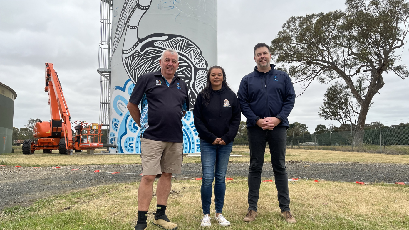 Three people stand in front of a grey water tower being painted. 