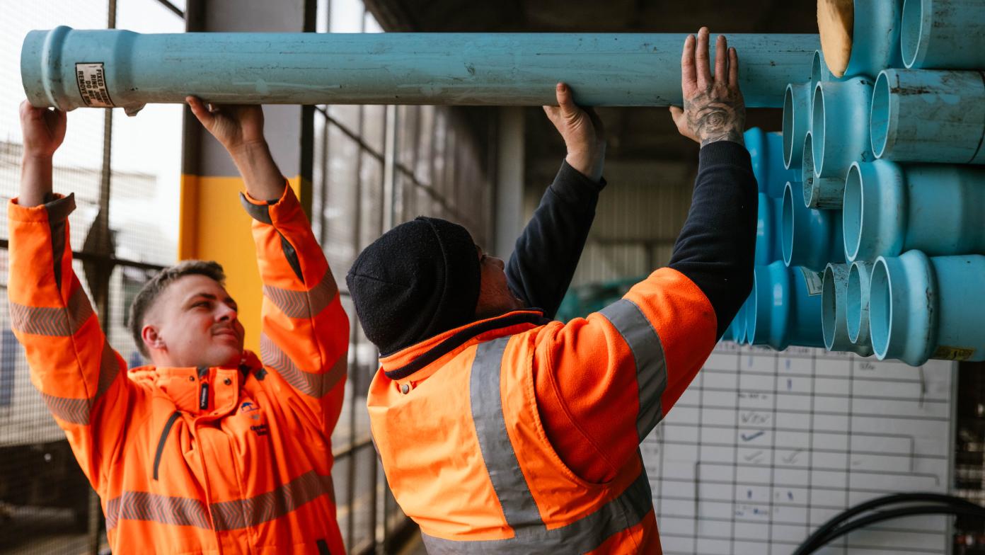 Two people in high visibility jackets remove blue pipes from the back of a  flatbed truck. 