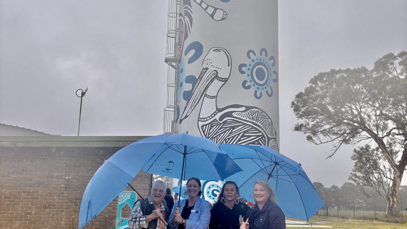 Four women stand in front of water tower with mural on it. They hold umbrellas. 