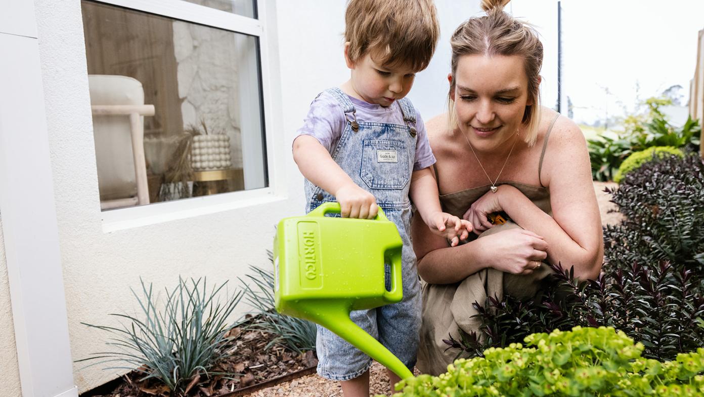 A woman and child water a garden with a little green watering can. 