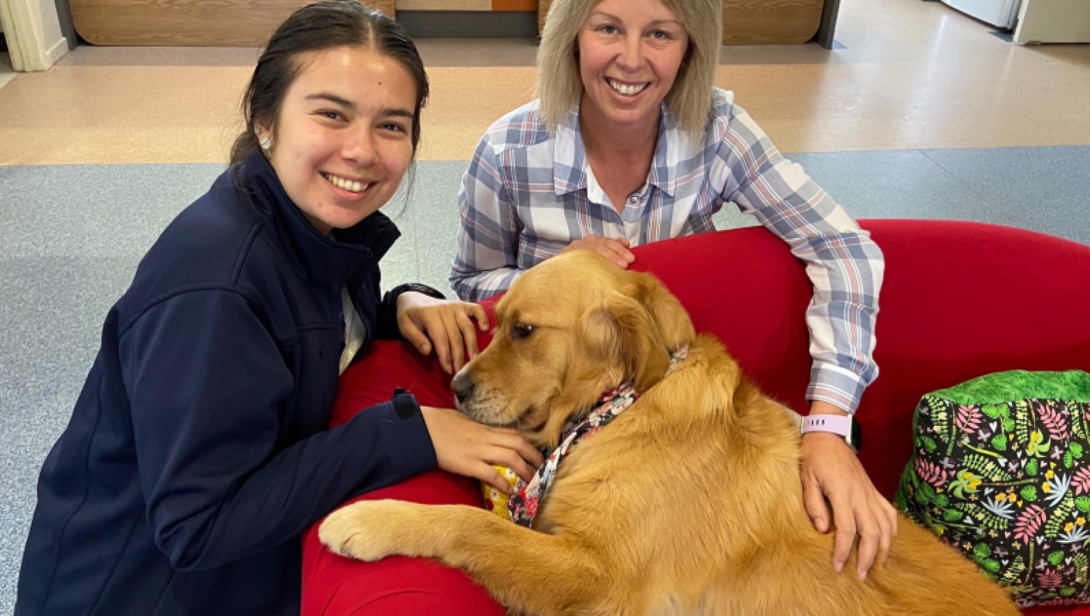 Two women smile while sitting with golden retriever. 