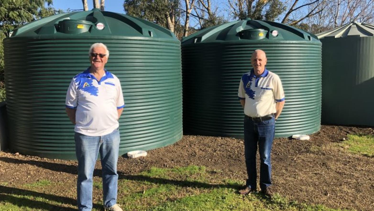 Two men stand in front of large green water tanks.