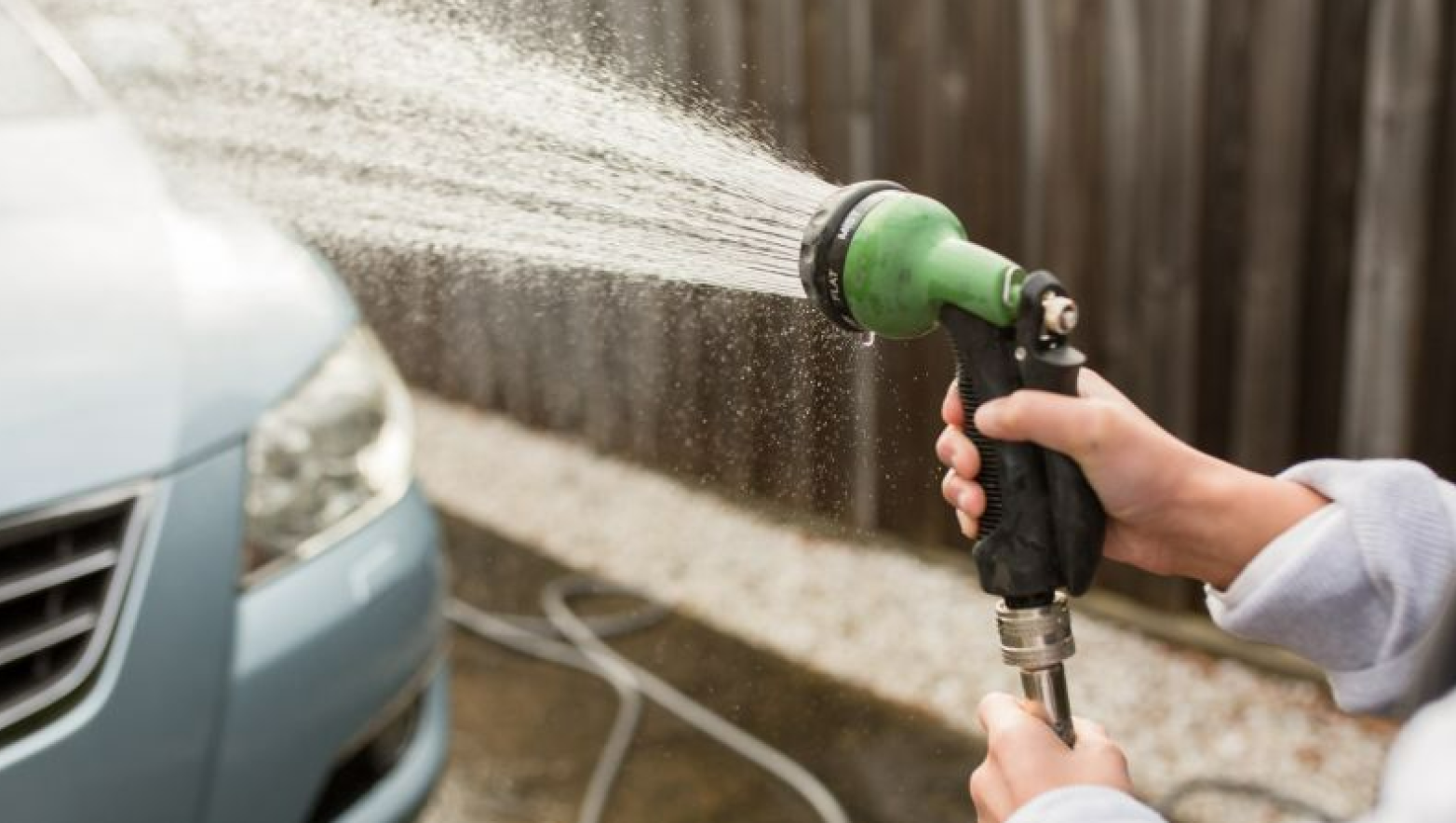 Water is being sprayed out of a hose at a light blue car. A person is holding the hose. 