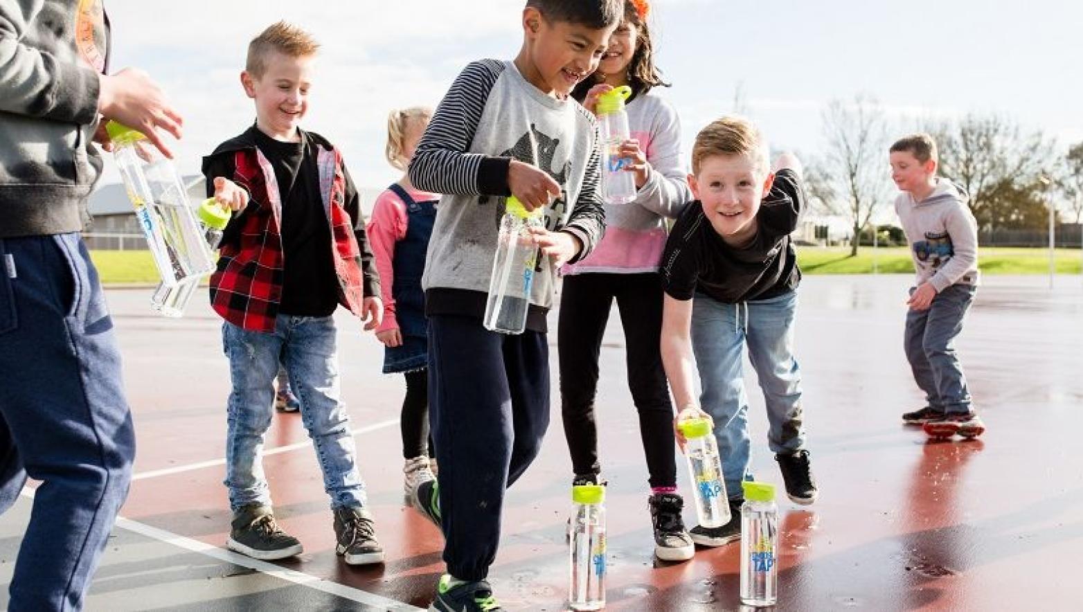 Children pick up water bottles off the ground of an orange netball court.