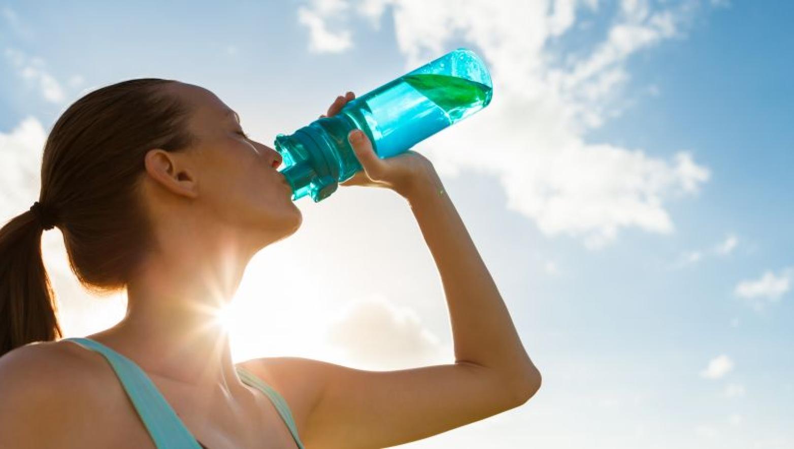 Sun shines behind woman drinking from a blue bottle of water.