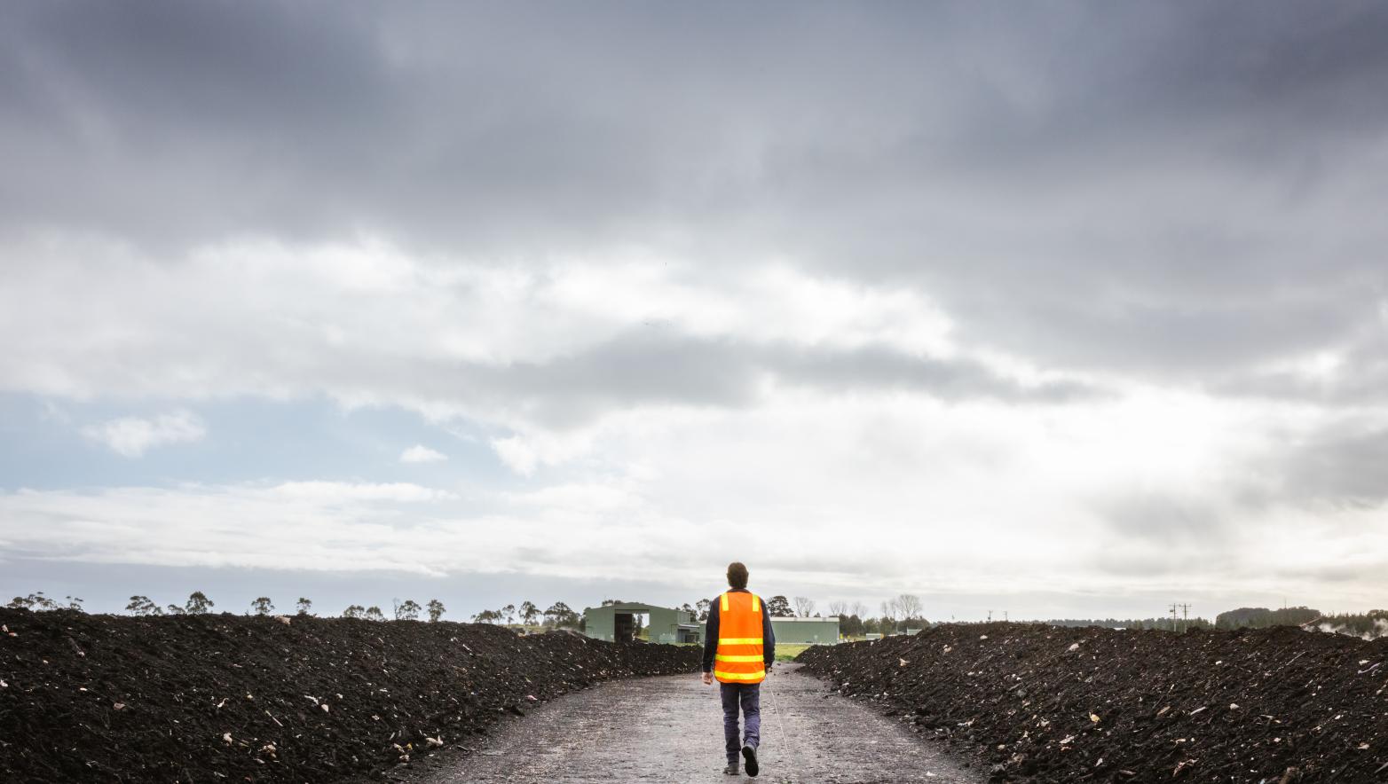 Person walks away from camera in high visibility vest, piles of compost are on either side of them.