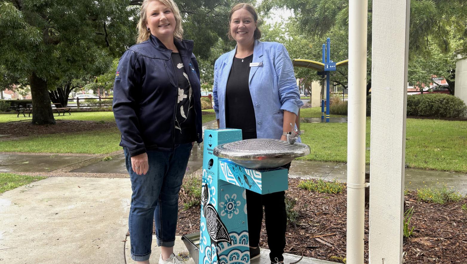 Two women stand behind a blue drinking fountain